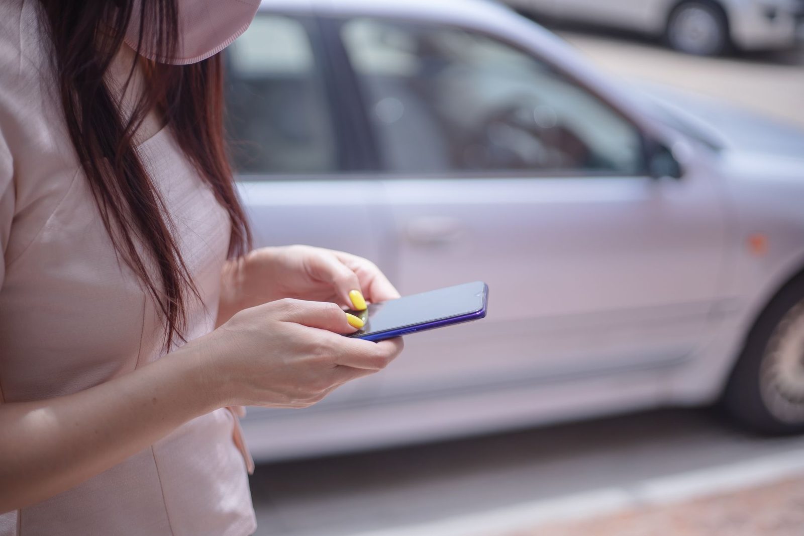 Mulher de máscara a usar um smartphone na rua, à frente de um carro cinzento estacionado