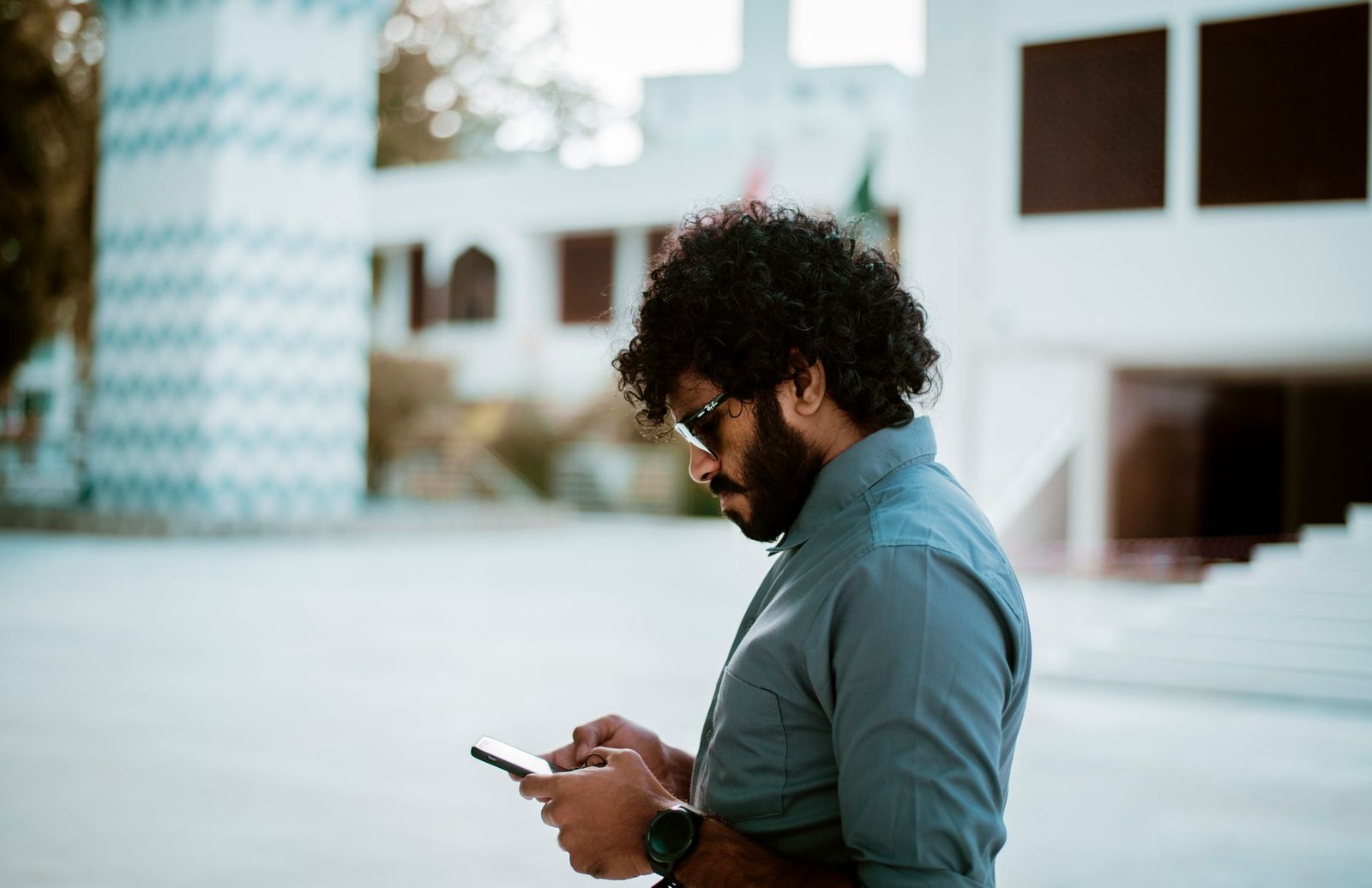 Bearded man wearing glasses checking his smartphone outdoors in front of a white building