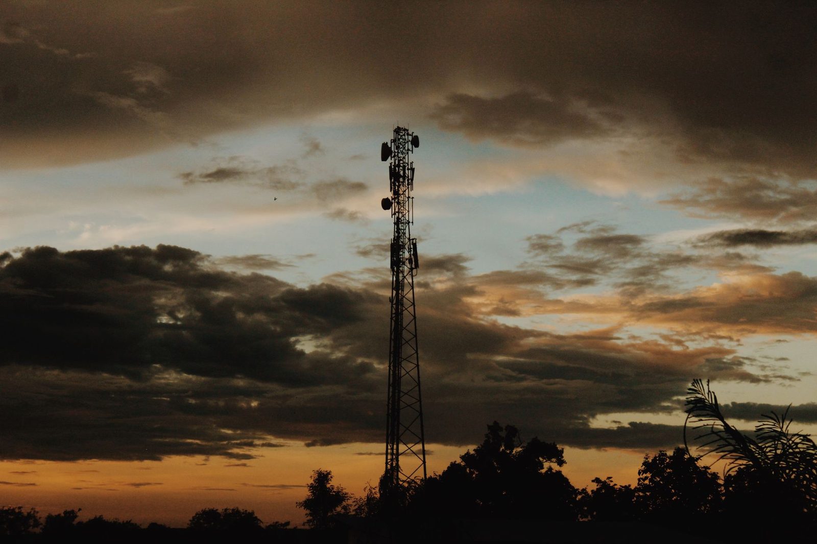 Maszt telefonii komórkowej z antenami, w kontrze na tle zachmurzonego nieba o zachodzie słońca