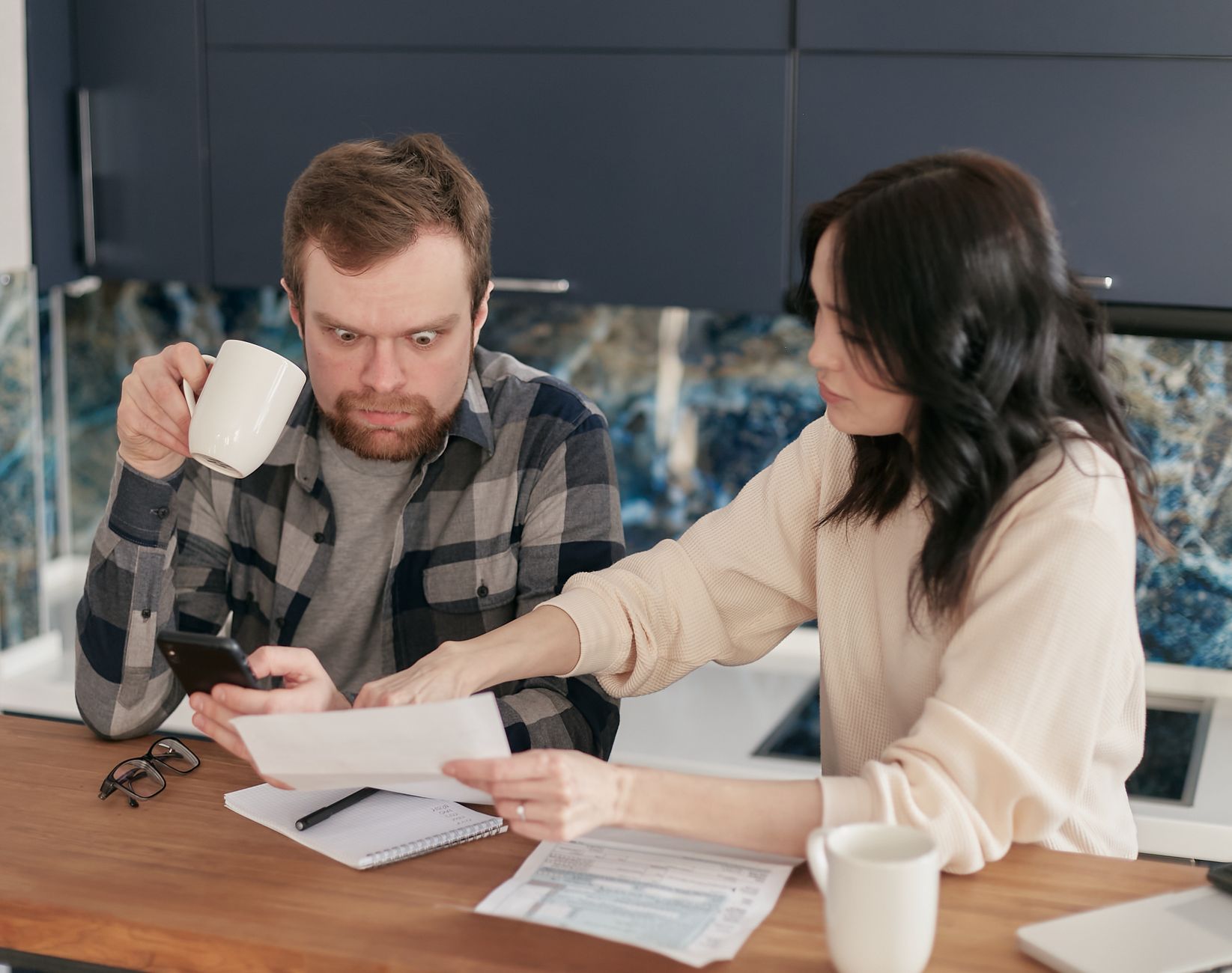 Un couple examine une facture papier et un smartphone dans une cuisine, l'homme tient une tasse