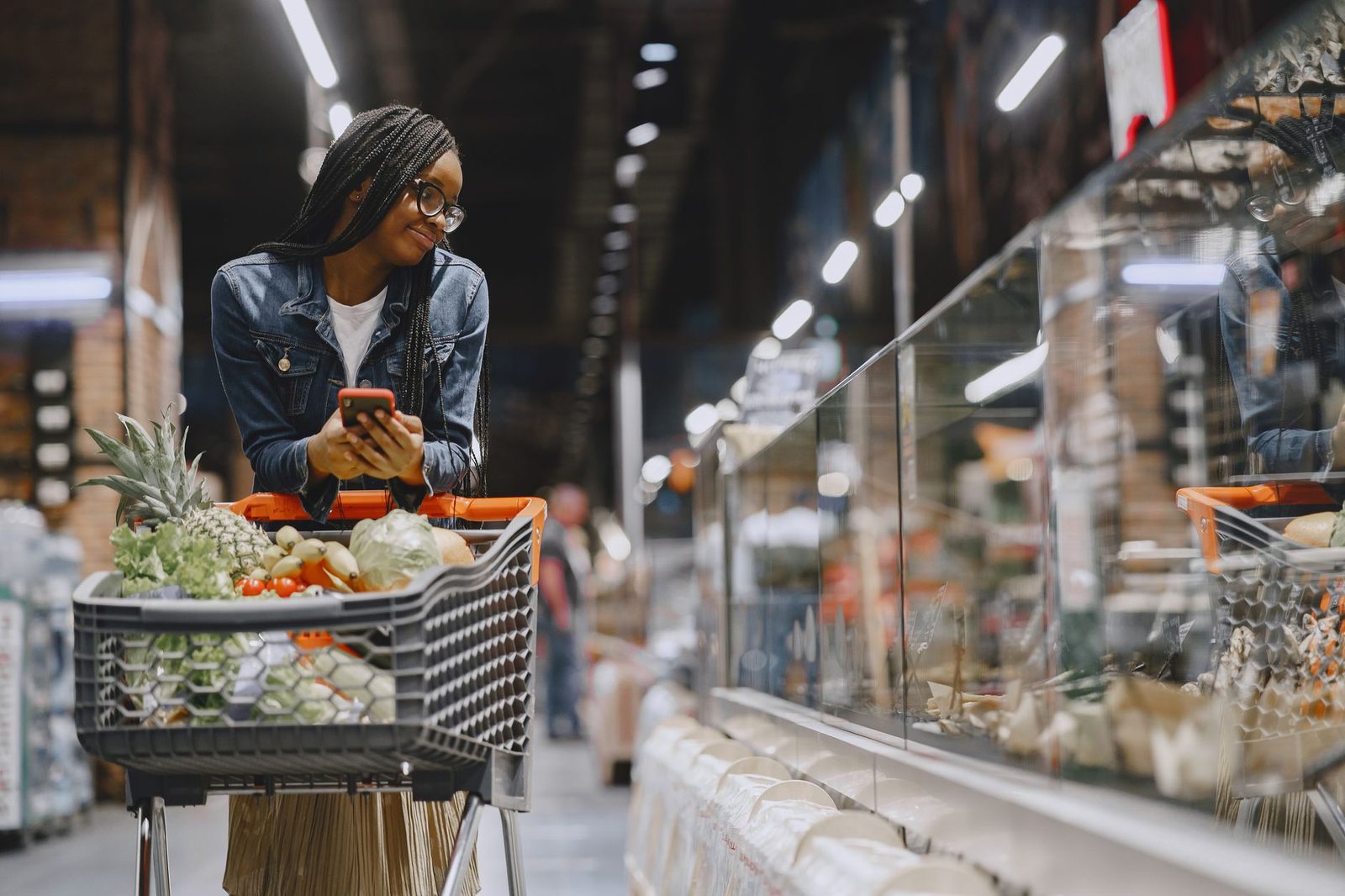 Vrouw met een winkelwagen vol groenten die haar smartphone raadpleegt in een supermarktschap