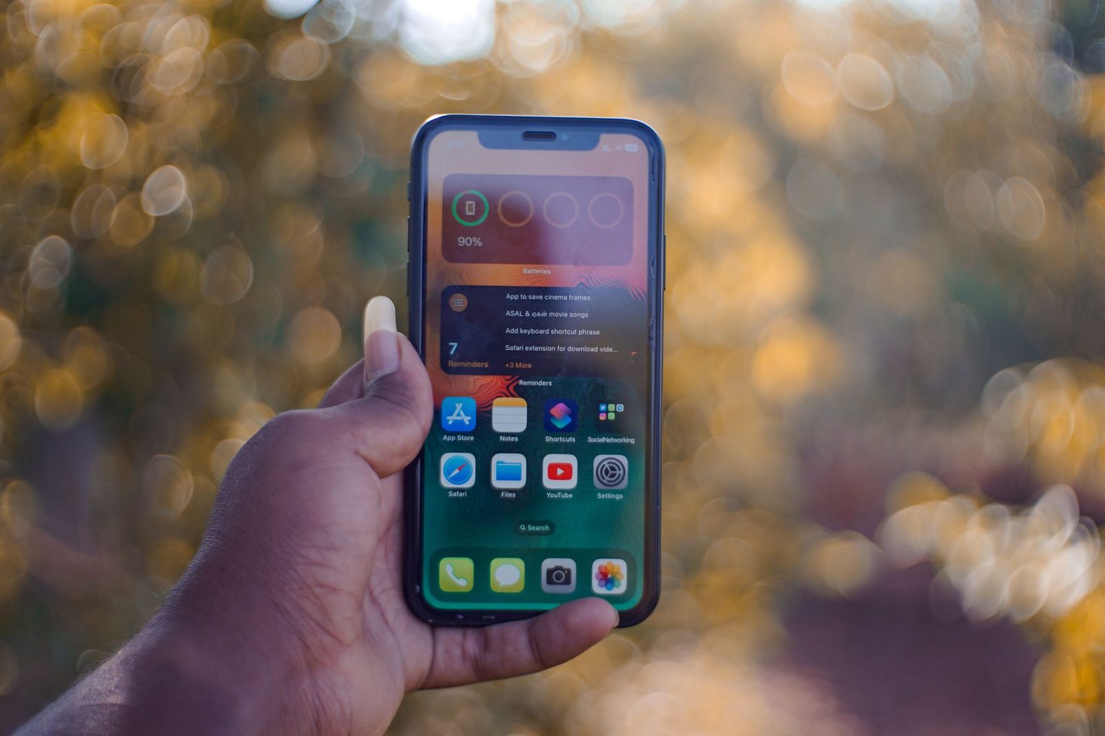 Mano che regge uno smartphone acceso con la schermata home, davanti a uno sfondo vegetale sfocato