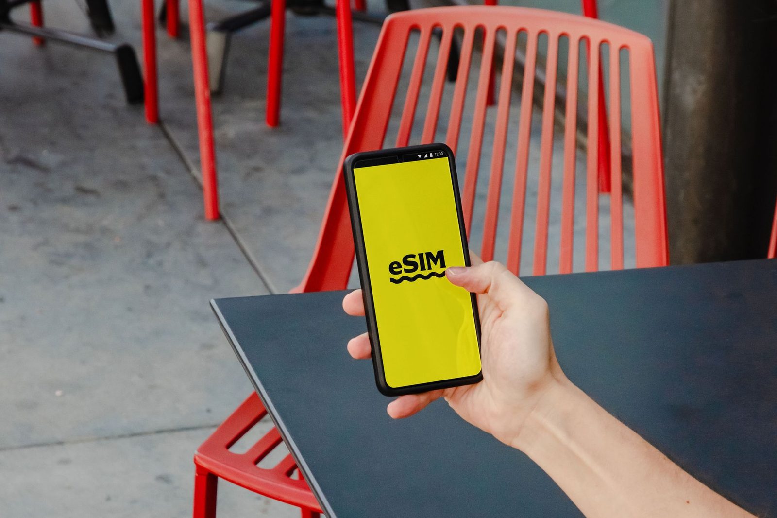 Hand holding a smartphone displaying "eSIM" on a yellow screen, in front of a table and red chairs
