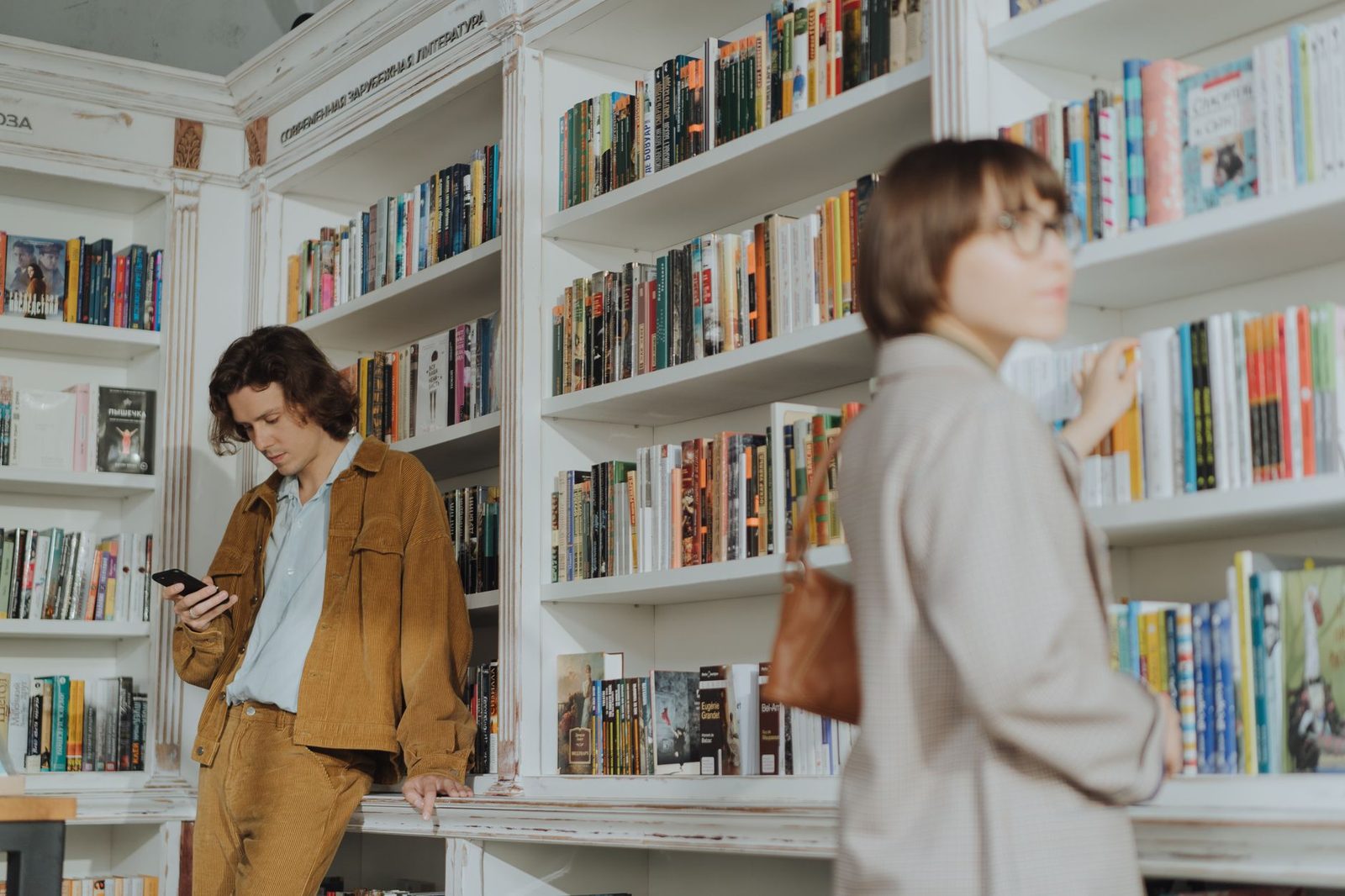 Jeune homme consultant son smartphone dans une librairie, une femme passant devant les étagères de livres