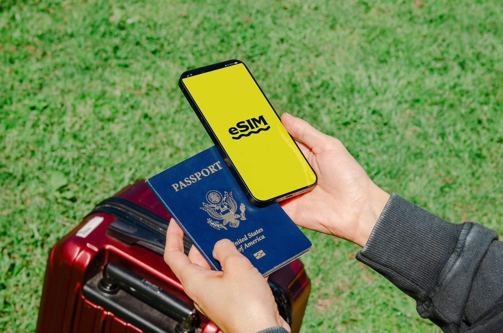 Mano che regge uno smartphone con la scritta «eSIM» e un passaporto statunitense sopra una valigia rossa
