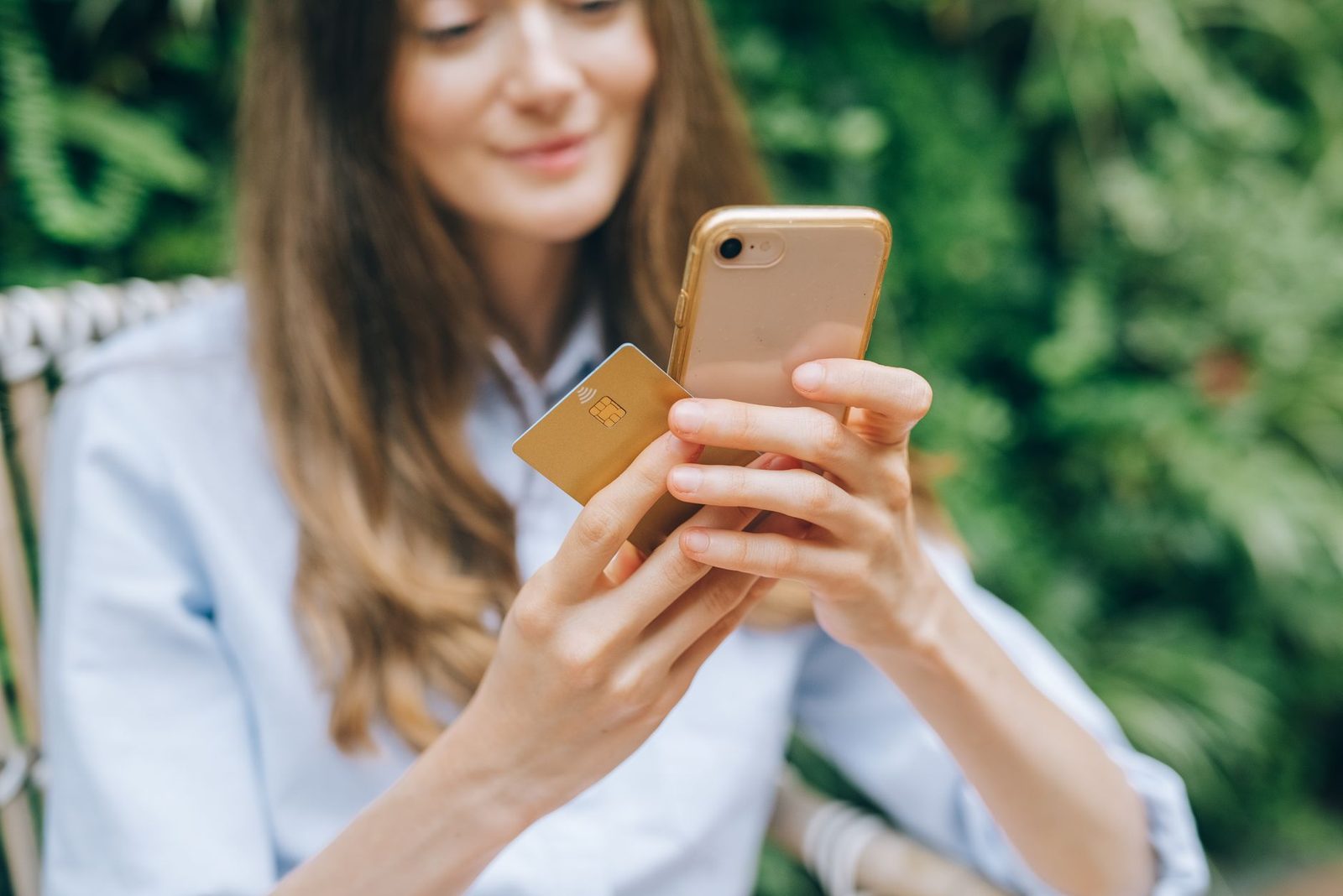 Junge Frau mit Smartphone und goldener Bankkarte, im Freien vor grünen Pflanzen sitzend