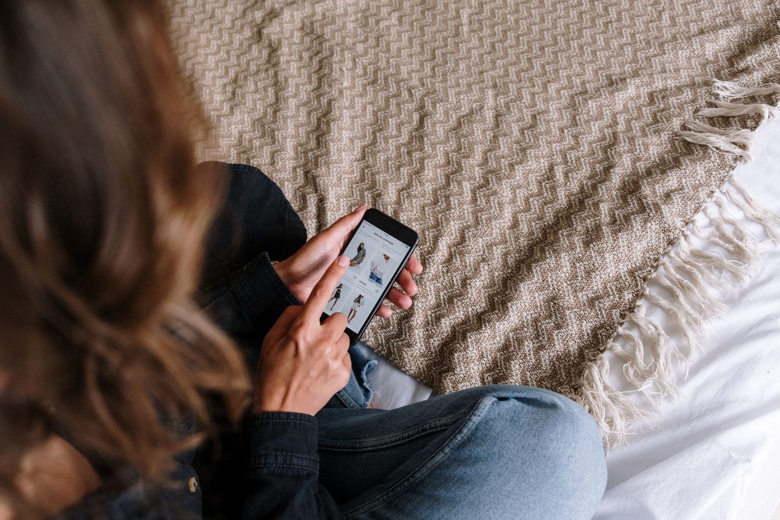 Donna seduta su un letto che usa uno smartphone tenuto con due mani, vista da dietro dall'alto