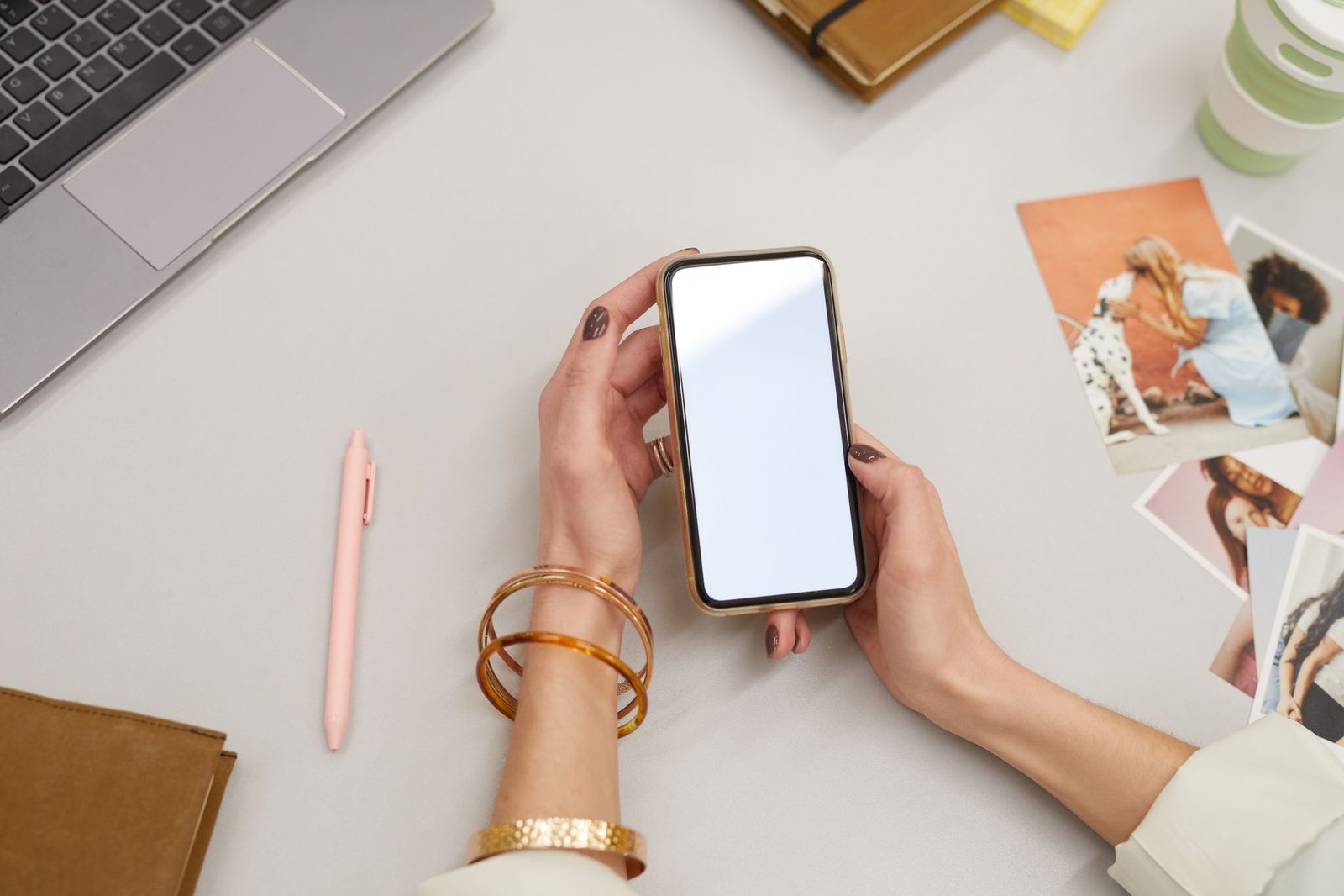Mains d'une femme tenant un smartphone à écran blanc au-dessus d'un bureau avec ordinateur portable et photos