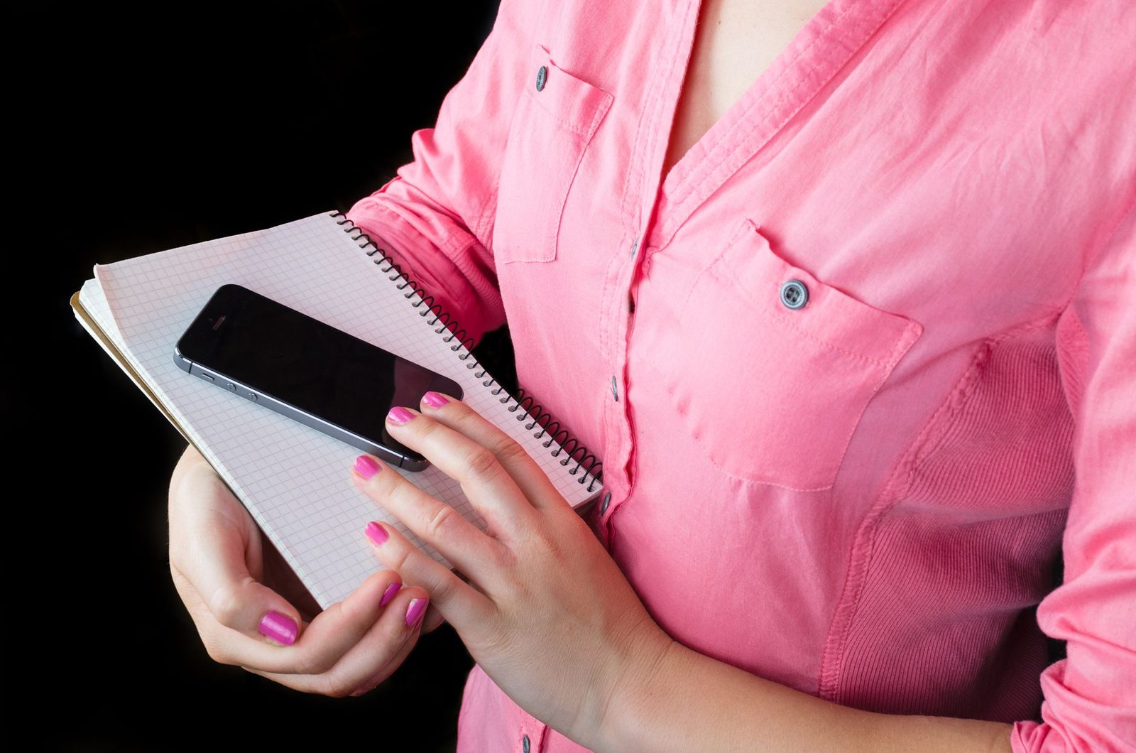 Mujer con camisa rosa sosteniendo un smartphone negro apoyado en un cuaderno de espiral, sobre fondo negro