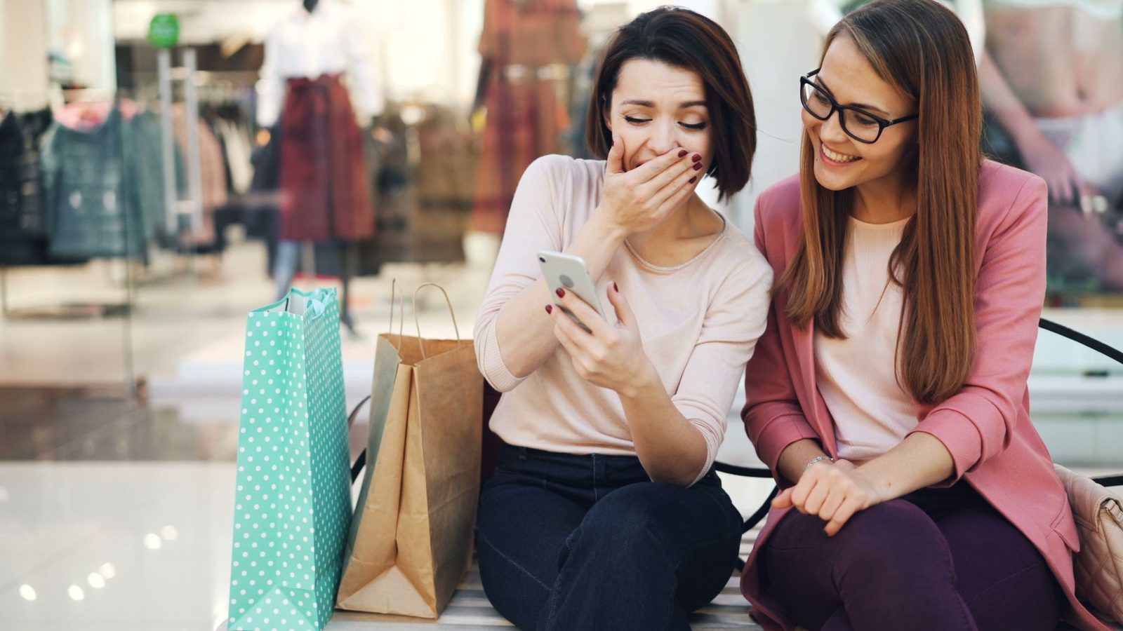 Deux jeunes femmes assises avec des sacs de shopping regardent ensemble l'écran d'un smartphone
