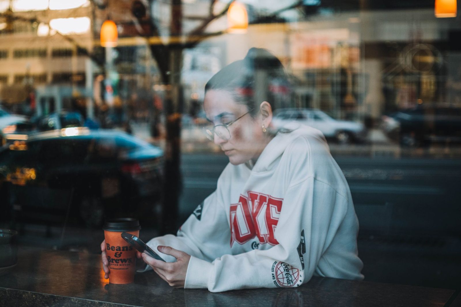 Jonge vrouw met bril die op haar smartphone kijkt achter het raam van een café, met een koffiebeker voor haar