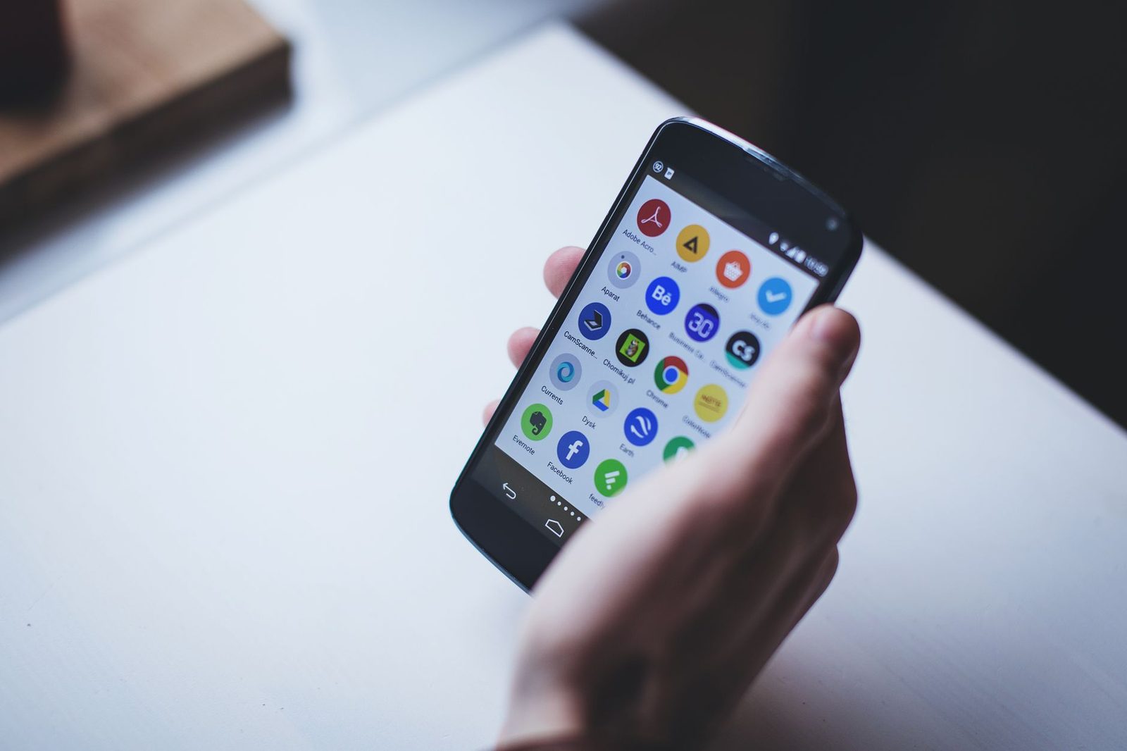 Hand met een smartphone waarop een raster van apps te zien is, boven een witte tafel