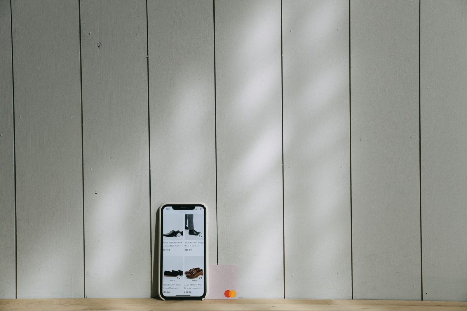Smartphone an einer hellen Holzwand, daneben eine Bankkarte, auf dem Display ein Online-Shop