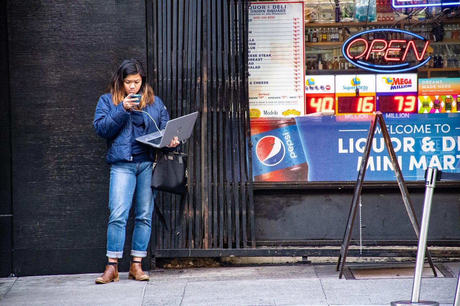 Frau auf der Straße, die auf ihr Smartphone schaut, einen aufgeklappten Laptop in der Hand