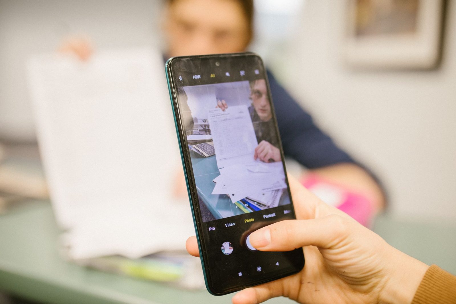 Hand hält ein Smartphone, dessen Display die Kamera zeigt, während Dokumente auf einem Tisch fotografiert werden
