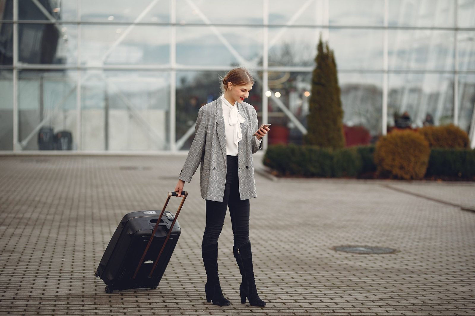 Woman with a suitcase checking her mobile phone in front of an airport's glass façade