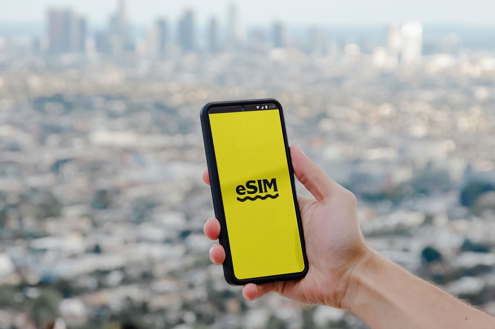 Hand holding a smartphone displaying "eSIM" on screen with a blurred city in the background