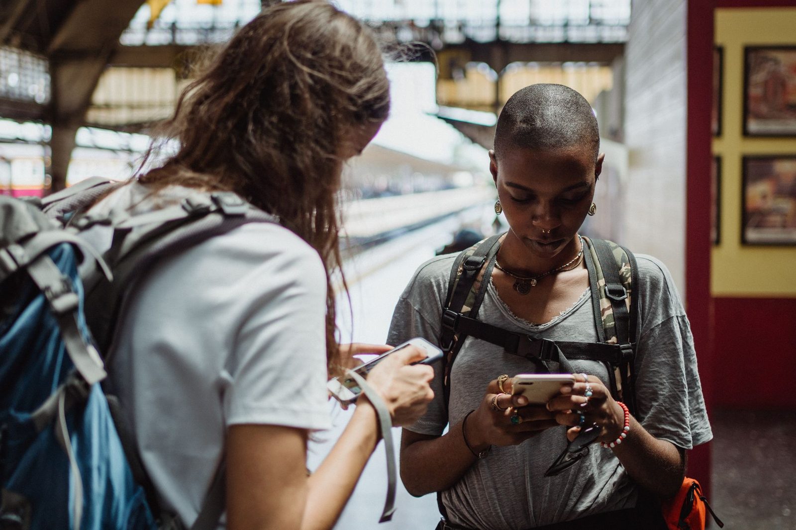 Two young women with backpacks checking their smartphones on a railway platform