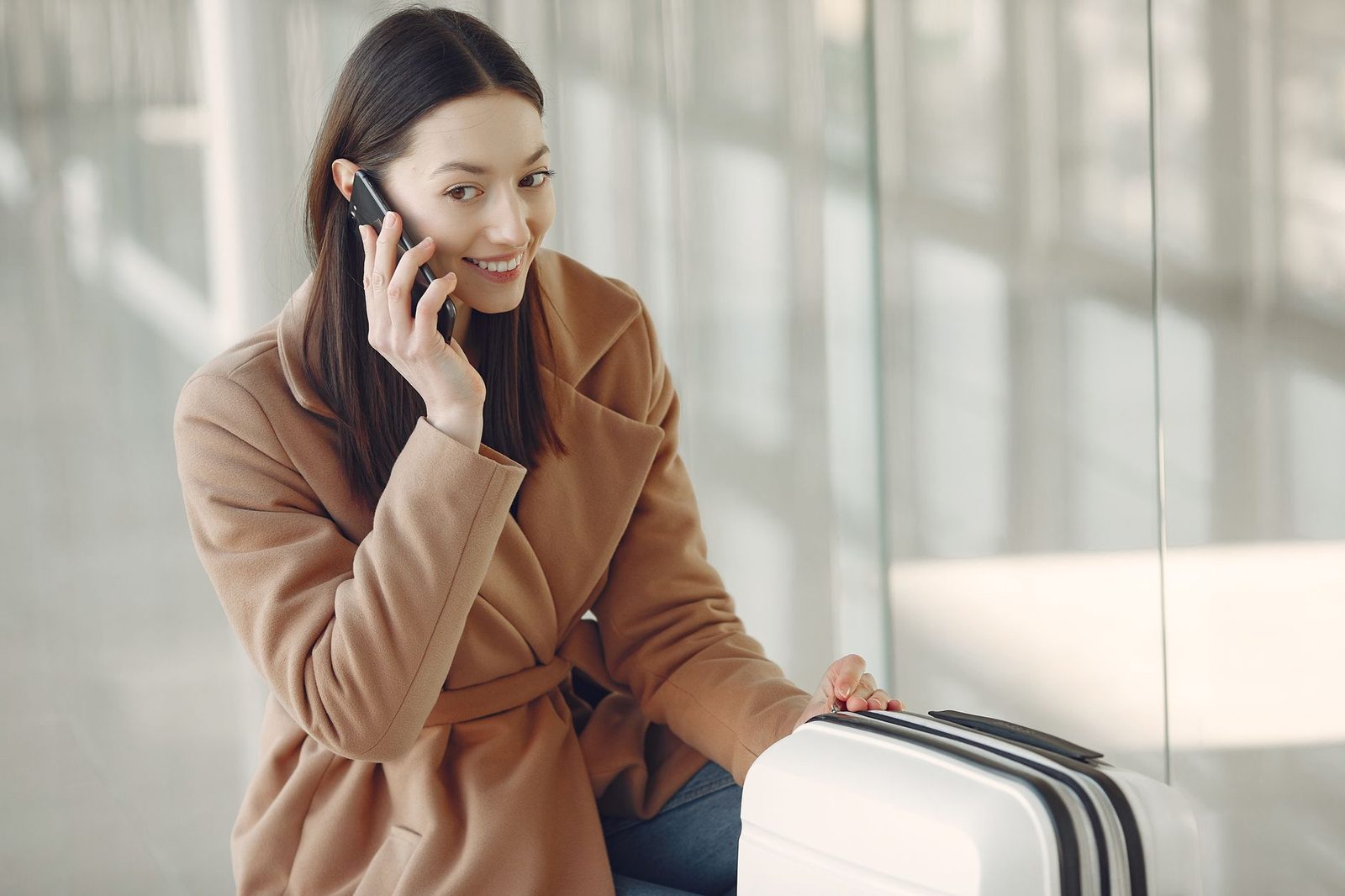 Woman in a beige coat talking on her smartphone, leaning on a white suitcase in an airport hall