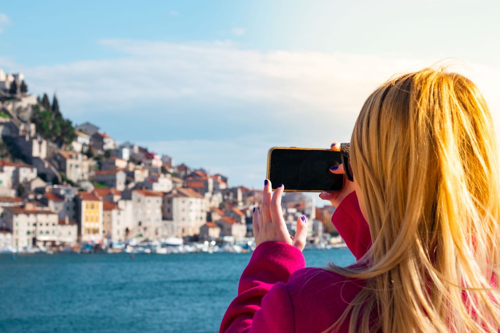 Blonde woman seen from behind photographing a Mediterranean coastal town by the water with her smartphone