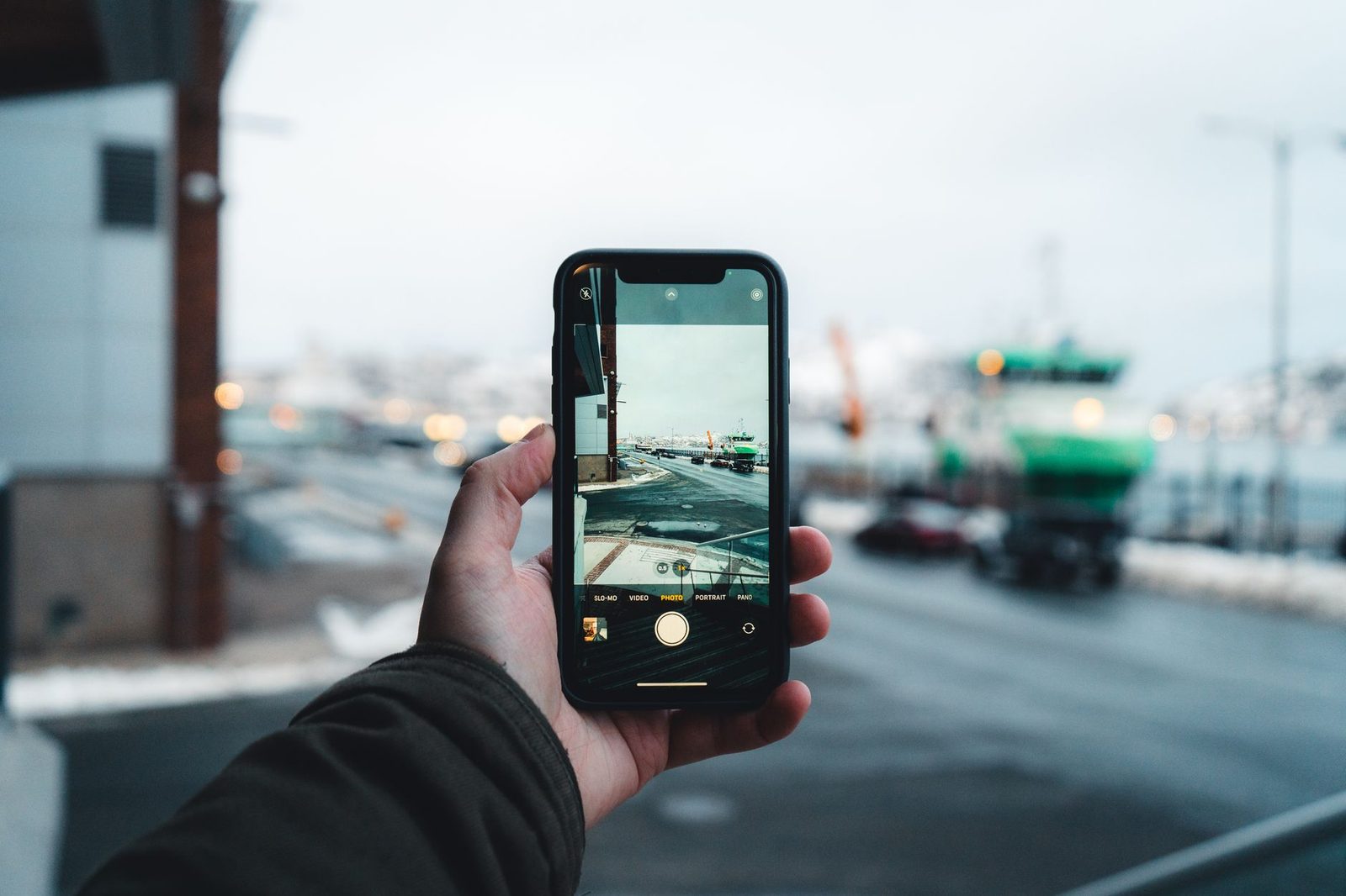 Hand holding a smartphone in camera mode in front of a snowy harbour with a green boat