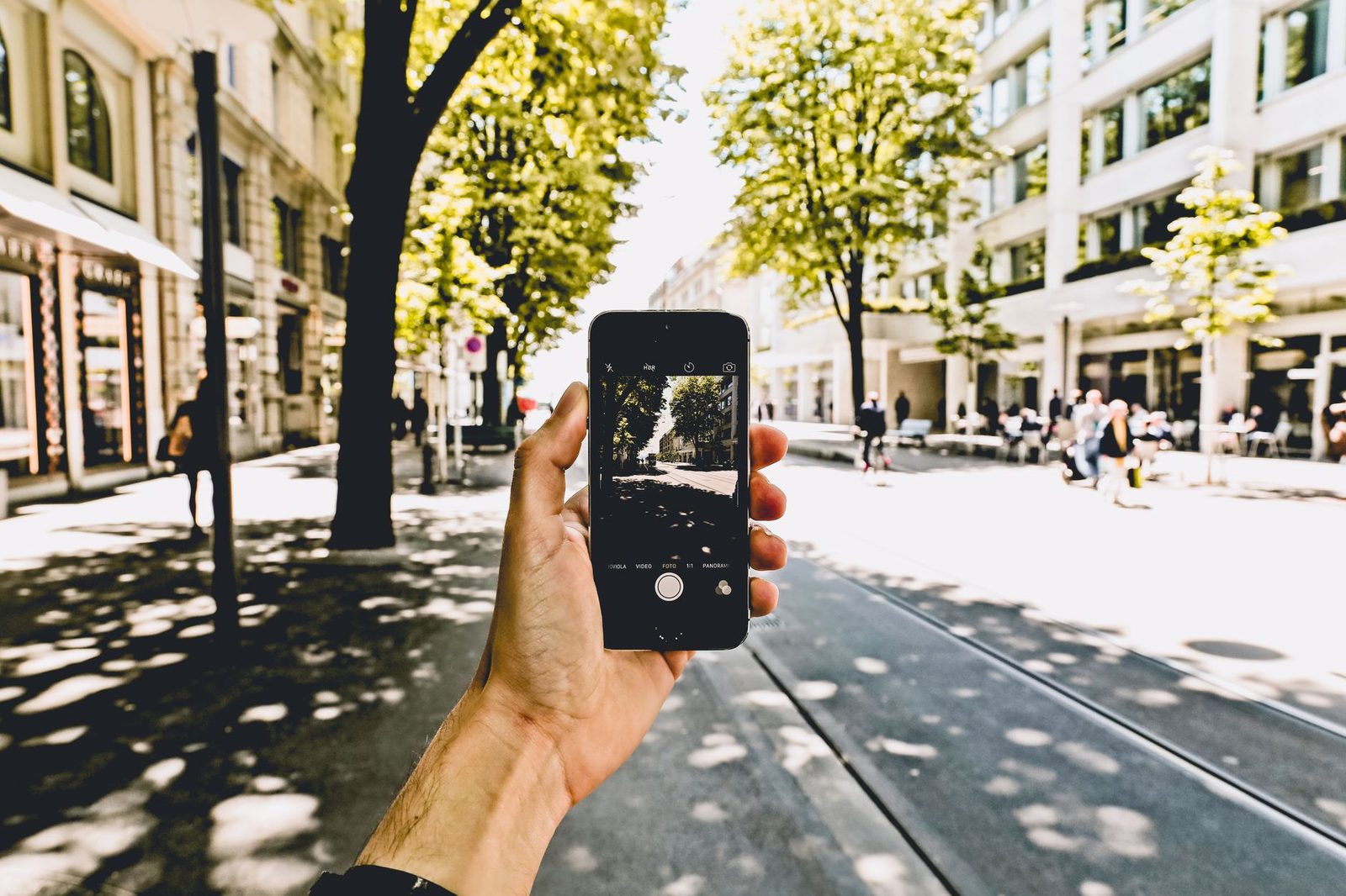 Hand holding a smartphone photographing a sunny tree-lined shopping street