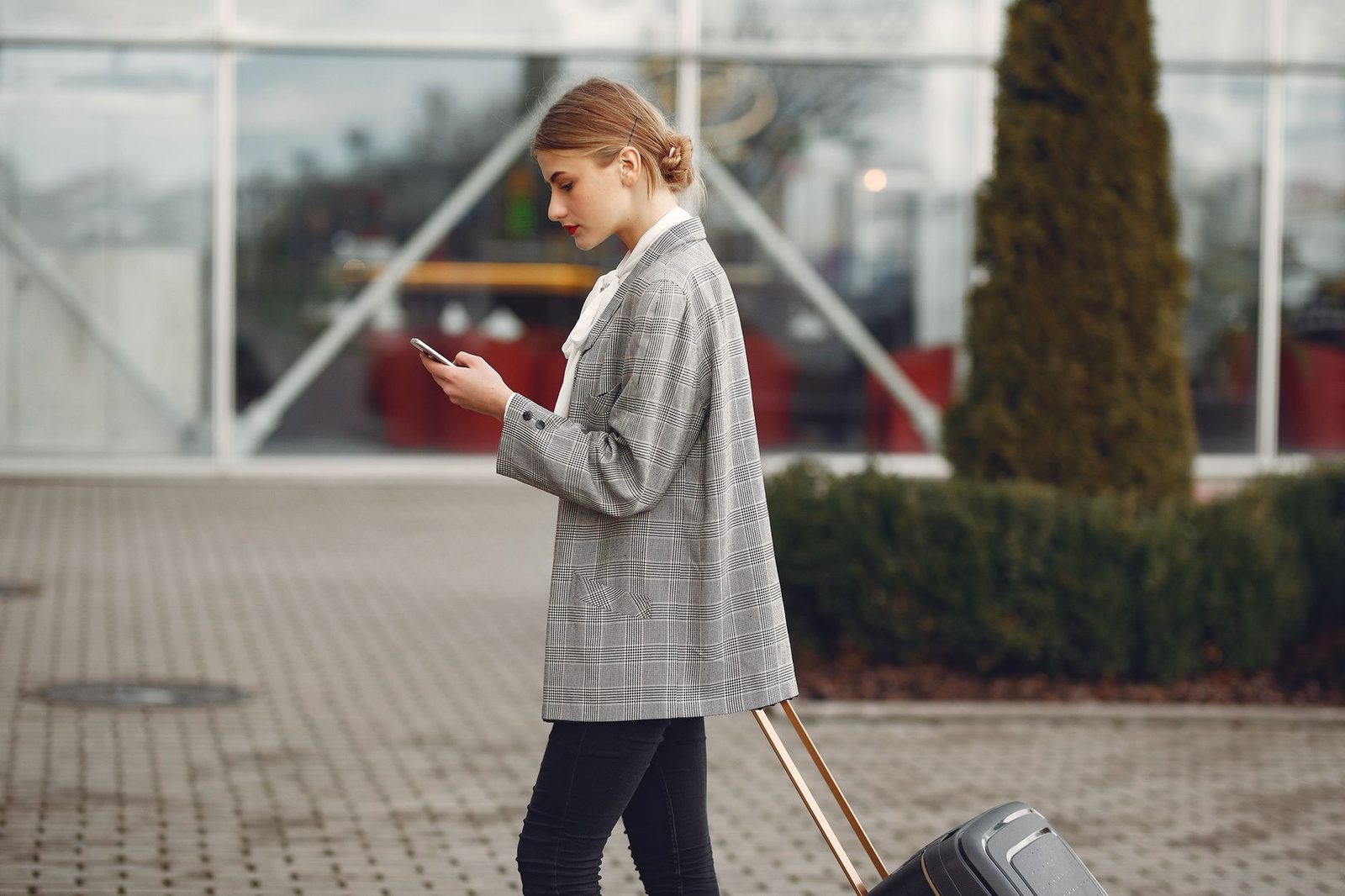 Woman in a checked jacket looking at her smartphone while pulling a suitcase outside an airport