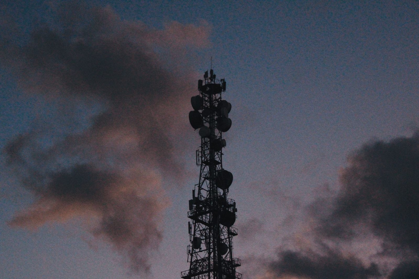 Mobile telecom tower covered in antennas, silhouetted against a cloudy sky at dusk